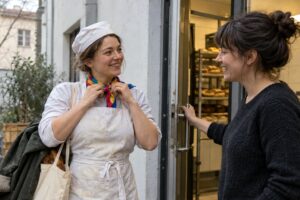 Frau in weißer Bäckerkleidung mit Mütze, mehlbestäubter Schürze und buntem Halstuch steht lächelnd vor einer Bäckerei, neben ihr eine Frau in schwarzem Pullover an der Ladentür
